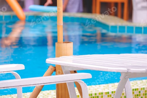 Preview: Empty white wooden table with chair and outdoor parasol on the edge of swimming pool