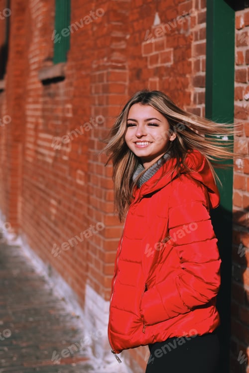 Preview: Young teenage girl in a red winter jacket walking alongside old orange brick wall
