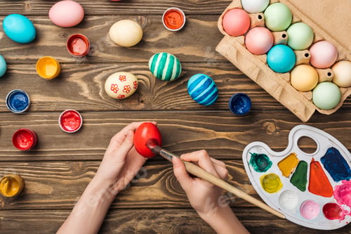 Preview: top view of girl painting easter eggs at wooden table with paints and color palette
