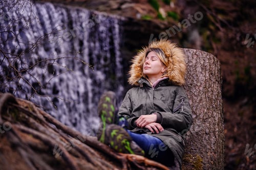 Preview: Young woman sits leaning on a stump in green parka with fur before waterfall alone in forest
