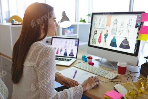 Preview: Side view of young Caucasian female fashion designer working on computer at desk in a modern office