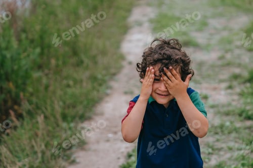 Preview: A playful little curly-haired boy covers his face with his hands, peeps, looks at camera at park