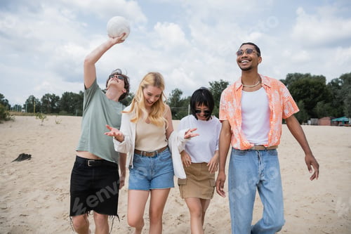 Preview: happy interracial friends walking on beach near man playing with ball