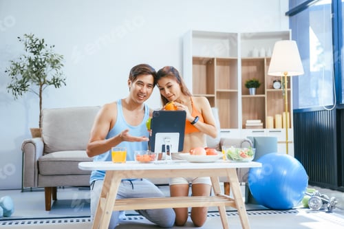 Preview: A happy young couple in sportswear sits on the sofa after exercising,enjoying fruits,salad,orange