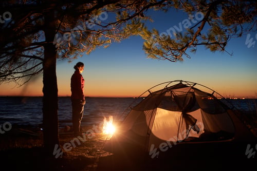 Preview: Woman having a rest at night camping near tourist tent, campfire on sea shore under starry sky