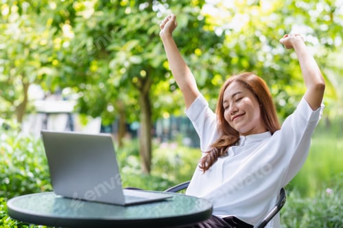 Preview: A woman with closed eyes, stretching arms and relaxing while working on laptop computer in the park