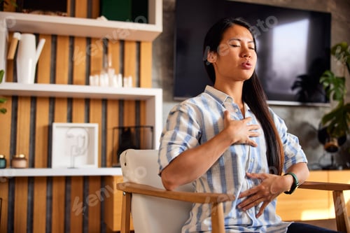 Preview: A relaxed Asian woman practicing yoga, sitting on an armchair at home.