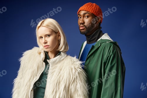 Preview: blonde woman posing with african american man in winter attire, trendy models on blue backdrop