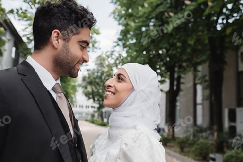 Preview: happy muslim bride in white hijab and groom in suit looking at each other