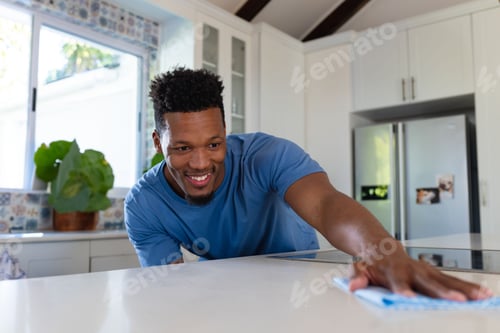 Preview: African American man leaning on home kitchen island wiping countertop with cloth under sunny window