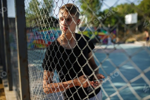 Preview: Cool boy in black T-shirt standing behind mesh fence and thoughtfully looking aside with cellphone