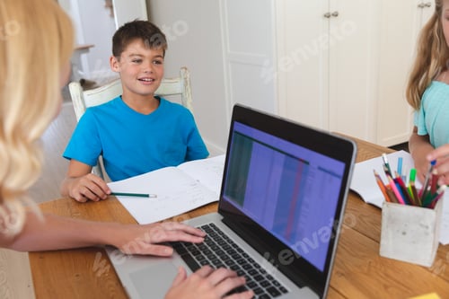 Preview: Caucasian mother using laptop and doing homework with her daughter and son smiling in kitchen