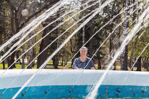 Preview: Senior woman enjoys looking at jets of water in fountain. In spring, city fountains begin their work