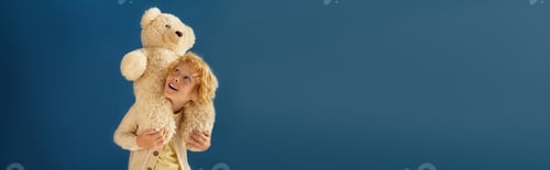 Preview: Blonde boy in casual clothes joyfully plays with teddy bear on blue background