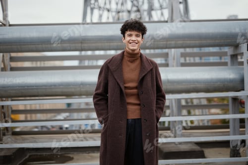 Preview: Fashionable young man in brown coat and turtle neck poses in front of a bridge, smiling for the