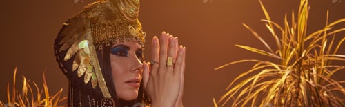 Preview: Woman in egyptian attire doing praying hands gesture near desert plants isolated on brown, banner