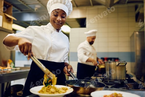Preview: Happy black female chef serving food while working in a restaurant kitchen.