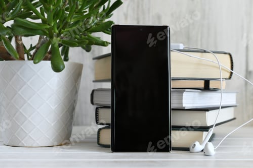 Preview: Phone with blank black screen for mock up against stack of books with earbuds