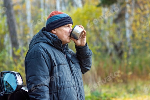 Preview: An elderly man drinks hot tea from a thermos near his car on the background of an autumn forest