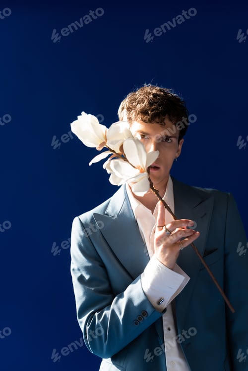 Preview: Stylish man in blazer holding magnolia flowers near face isolated on navy blue