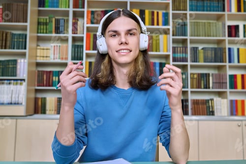 Preview: Teenage college student in headphones looking at webcam, sitting at desk in library