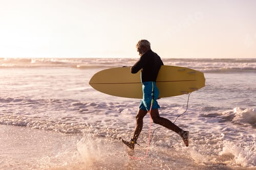 Preview: Senior African American man in swim shorts carrying yellow surfboard wading on beach at sunset