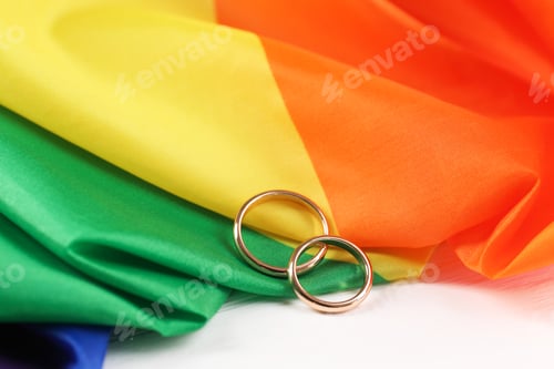 Preview: Rainbow LGBT flag and wedding rings on white wooden table, closeup