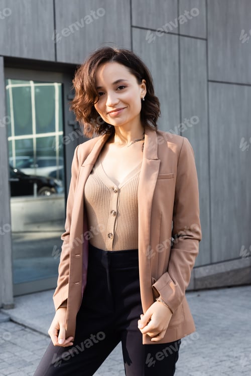 Preview: brunette woman in beige blazer smiling at camera while standing outdoors