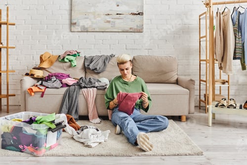 Preview: young tattooed woman in casual clothes sorting thrift store finds while sitting on floor near couch