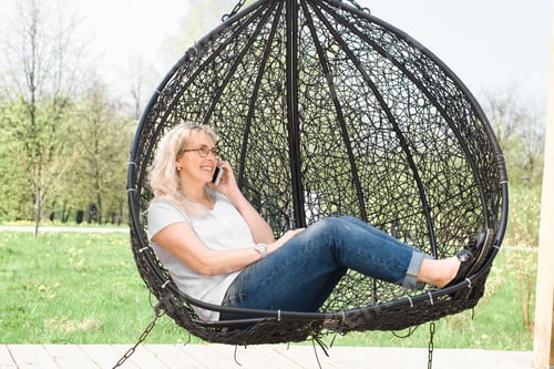Preview: Happy woman talking on a cell phone and sitting in a hanging swing in a summer park. Smiling lady