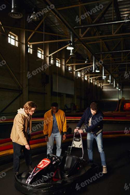 Preview: happy interracial friends standing near red go-kart inside of indoor track, motorsport hobby