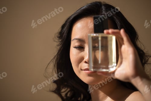 Visualização: Mulher asiática segurando um frasco de perfume perto do rosto, mostrando a fragrância