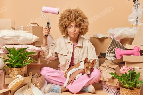 Preview: Irritated curly woman has much work to do holds paint roller going to refurbish walls in room poses
