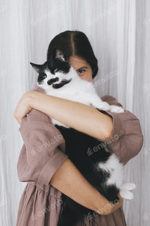 Preview: Stylish happy woman in linen dress hugging funny cat on background of pastel fabric. Pet and friend