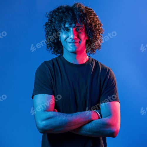 Preview: Posing for a camera. Young beautiful man with curly hair is indoors in the studio with neon lighting