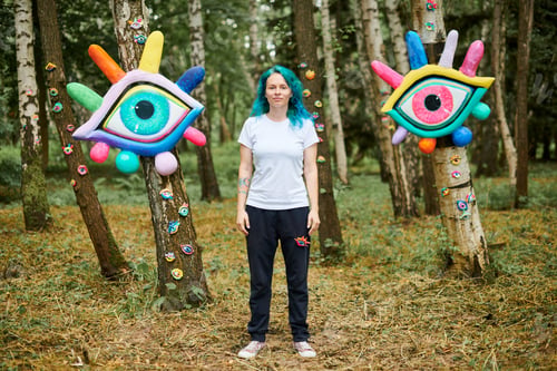 Preview: Weird young adult woman with dyed turquoise hair in white T shirt near big eyes art installation