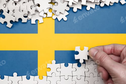 Preview: Sweden flag is depicted on a table on which the human hand folds a puzzle of white color.