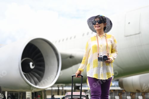 Preview: Happy 60s woman in summer clothing and sunglasses walking near the airport with a suitcase.