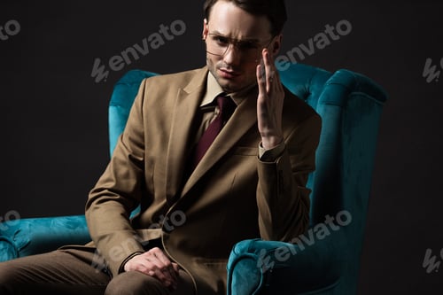 Preview: pensive elegant man in beige suit eyeglasses sitting in blue velour armchair isolated on black
