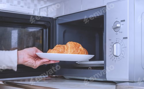 Preview: Woman hand putting plate of croissants into microwave oven in the kitchen