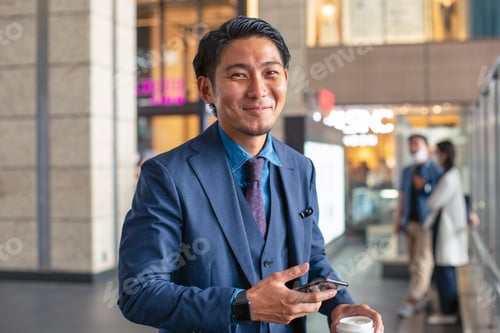 Preview: A young businessman in a blue suit on the move in a city downtown area, holding his mobile phone.