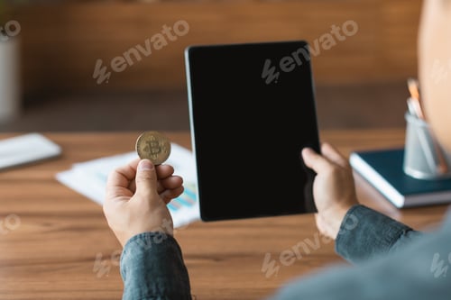 Preview: Adult asian guy looks at tablet with empty screen and bitcoin coin, analyzes financial data at