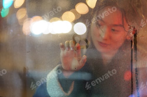 Preview: Cute chinese girl paints on foggy glass window in heart at night, enjoying rainy weather at autumn.
