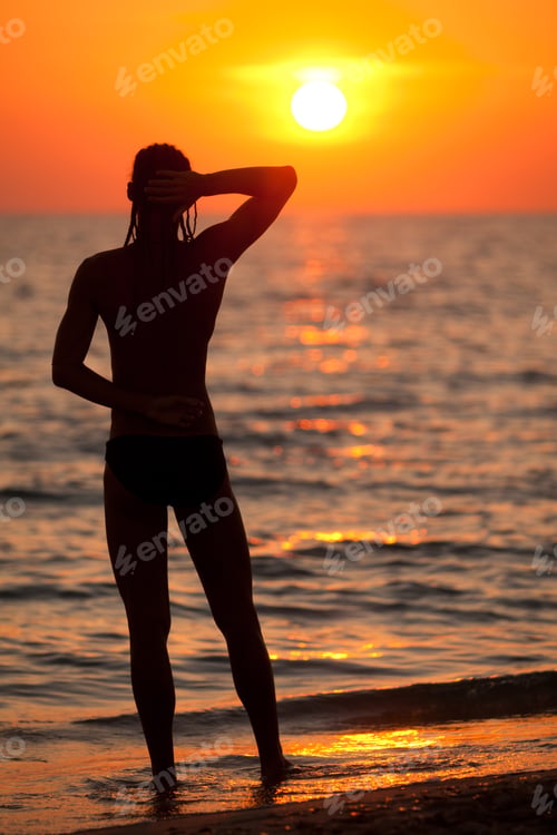 Preview: Silhouette of man standing backwards at sea water edge and enjoying sunset