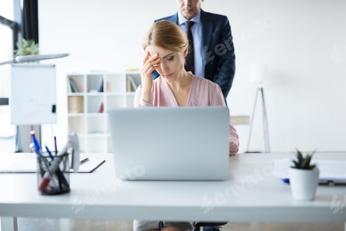 Preview: pensive businesswoman with laptop and documents working in office, businessman standing behind
