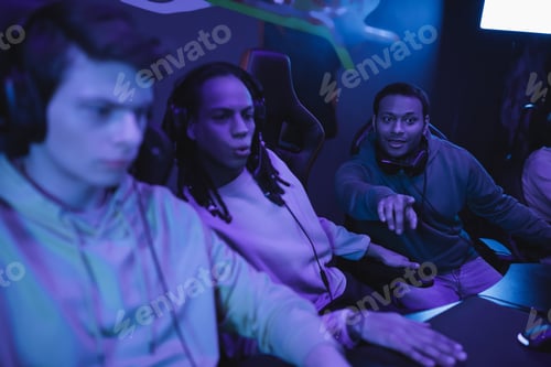 Preview: Indian man pointing with finger near interracial friends in gaming club