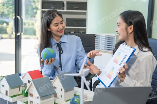 Preview: Two businesswomen sit at a table in the office,discussing a model village with windmills