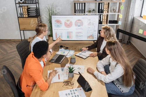 Preview: Group of multiracial female financial analysts sitting together at office desk and examining graphs