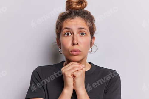 Preview: Horizontal shot of pretty young European woman with hair bun keeps hands under chin stands