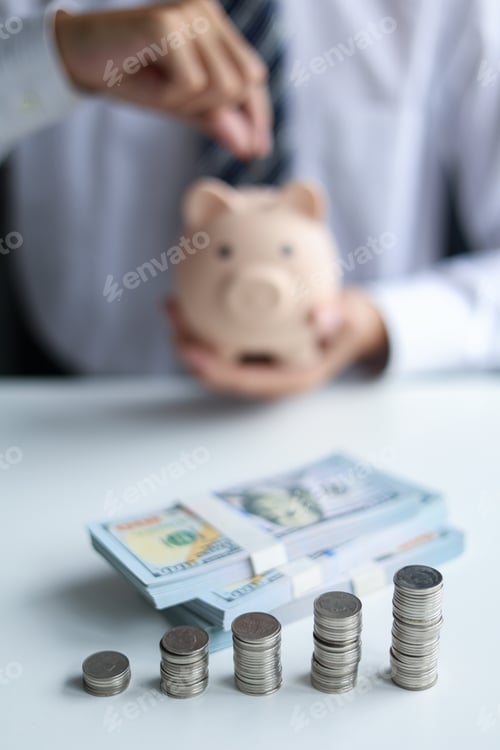 Preview: Stacks of coins and money on the table with businessman putting coin in piggy bank at the back.